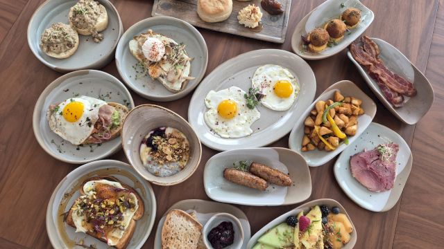 A spread of brunch plates: sunny-side-up eggs, yogurt with granola, toasted bread, pastries, fruit, sausages, bacon bits, croissants, nuts, berries, and small veggie plates, all on gray platters.
