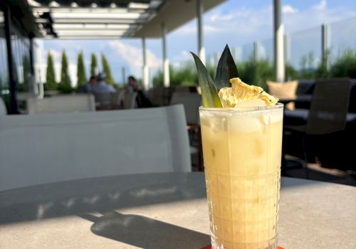 A tall iced cocktail in a faceted glass with citrus and mint sits on an outdoor patio with people and greenery in the background.
