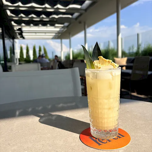 A tall iced cocktail in a faceted glass with citrus and mint sits on an outdoor patio with people and greenery in the background.