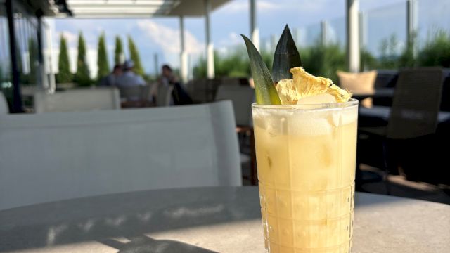 A tall iced cocktail in a faceted glass with citrus and mint sits on an outdoor patio with people and greenery in the background.