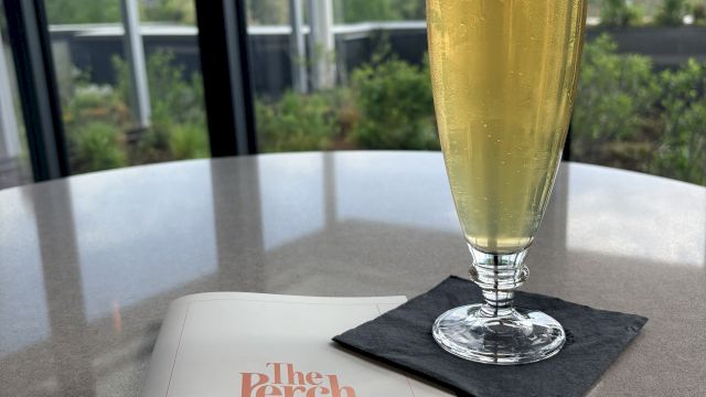 A tall flute of champagne on a napkin beside an open menu on a patio table, with a sunny outdoor view in the background.