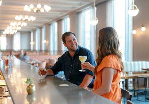 Two people smile and toast with drinks in a bright, modern bar/restaurant with pendant lights.