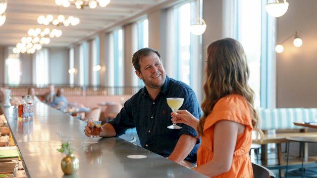 Two people smile and toast with drinks in a bright, modern bar/restaurant with pendant lights.