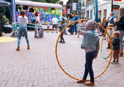 A girl with a large hoop stands in a lively outdoor scene, others walk and play in a brick-paved square with tents and colorful banners.