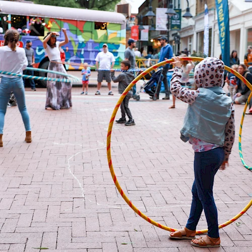 A girl with a large hoop stands in a lively outdoor scene, others walk and play in a brick-paved square with tents and colorful banners.