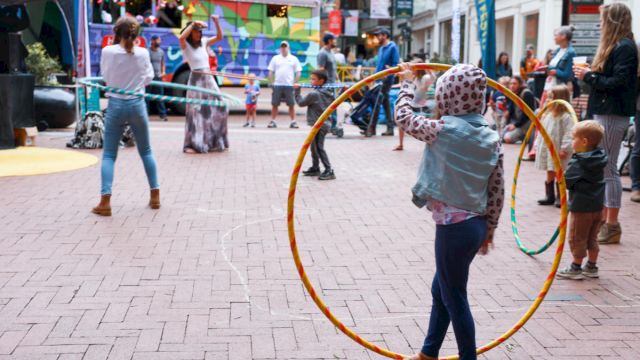 A girl with a large hoop stands in a lively outdoor scene, others walk and play in a brick-paved square with tents and colorful banners.