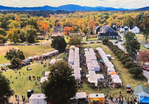 Aerial view of a festival with long row tents, crowds on the lawns, fall foliage, and houses in the background under a blue sky.