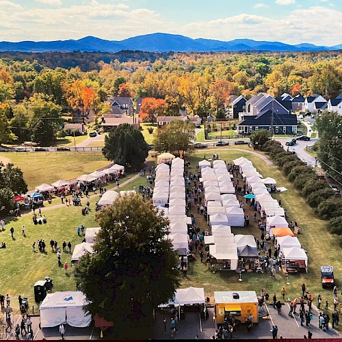 Aerial view of a festival with long row tents, crowds on the lawns, fall foliage, and houses in the background under a blue sky.