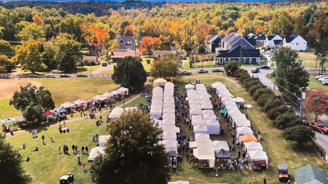 Aerial view of a festival with long row tents, crowds on the lawns, fall foliage, and houses in the background under a blue sky.