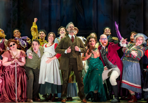 A lively theater cast in colorful 19th-century costumes celebrates on stage, singing and posing with joyful, theatrical energy.