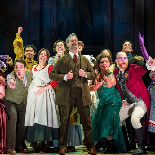 A lively theater cast in colorful 19th-century costumes celebrates on stage, singing and posing with joyful, theatrical energy.