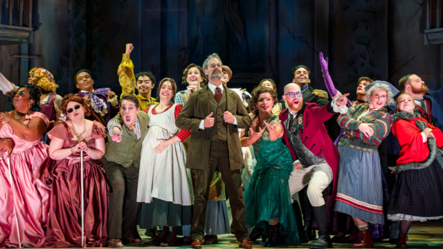 A lively theater cast in colorful 19th-century costumes celebrates on stage, singing and posing with joyful, theatrical energy.