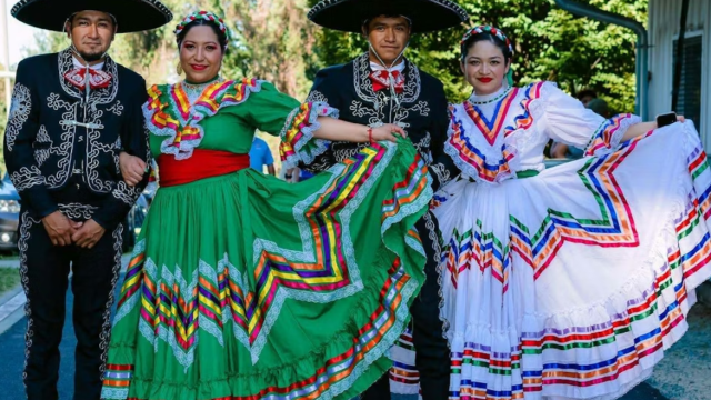 A group of four people wearing colorful traditional Mexican folkloric dresses and charro outfits, posing outdoors with a bright backdrop.