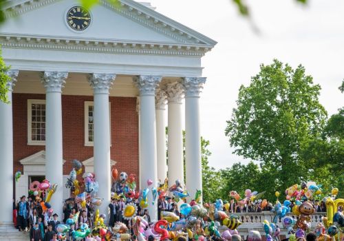 A festive crowd on the steps of a red-brick neoclassical building, balloons and people gathering for a celebration in a sunny, leafy campus-like setting.