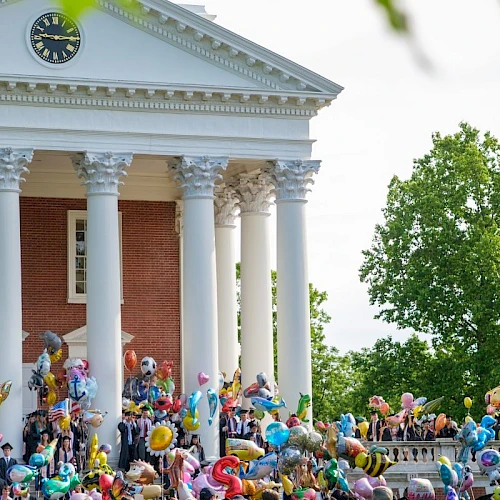 A festive crowd on the steps of a red-brick neoclassical building, balloons and people gathering for a celebration in a sunny, leafy campus-like setting.