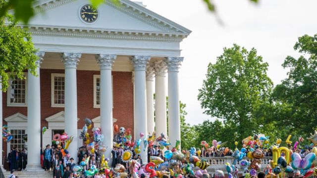 A festive crowd on the steps of a red-brick neoclassical building, balloons and people gathering for a celebration in a sunny, leafy campus-like setting.