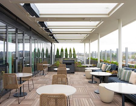 Rooftop terrace with wicker chairs, round white tables, and cushioned seating under a skylit canopy, overlooking a cityscape&mdash;bright and airy.