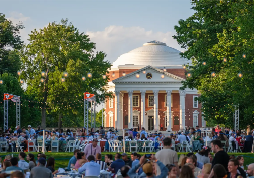 A large outdoor gathering in front of a domed neoclassical building, with people seated on chairs and string lights overhead, during the day.