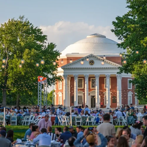 A large outdoor gathering in front of a domed neoclassical building, with people seated on chairs and string lights overhead, during the day.