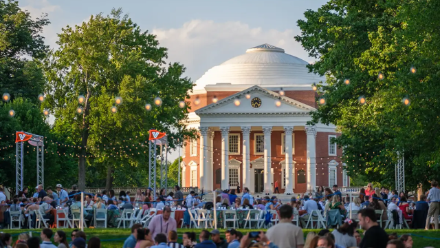 A large outdoor gathering in front of a domed neoclassical building, with people seated on chairs and string lights overhead, during the day.
