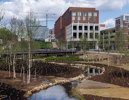 A modern campus scene with a small stream, trees, and contemporary brick buildings in the background, under a partly cloudy sky.