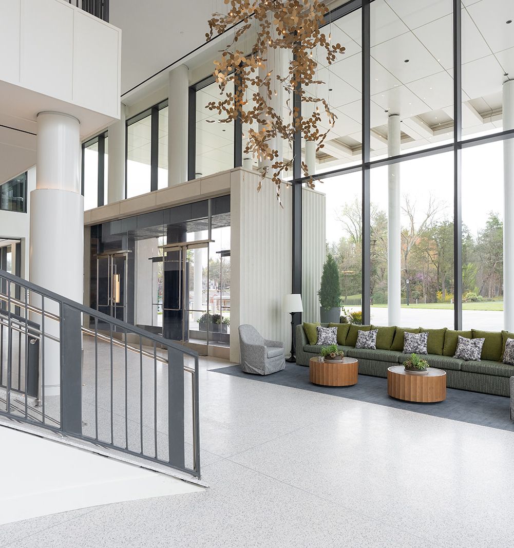 A bright, modern lobby with large glass walls, a staircase, and a seating area featuring green cushions and potted plants near a sunny outdoor view.