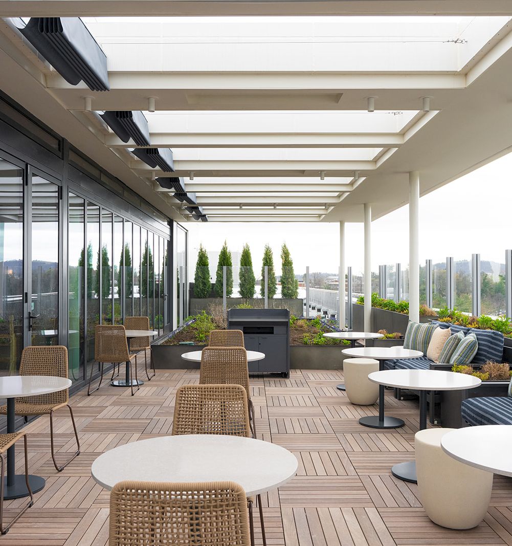 A modern outdoor rooftop terrace with glass walls, wicker chairs, white round tables, and a long sofa area under a skylight, overlooking the city.