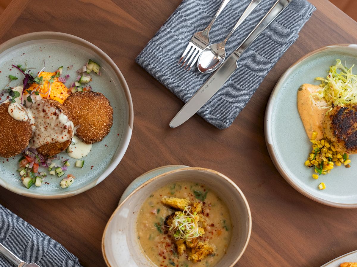 Atop view of three plates with plated dishes: croquettes or fried patties, garnishes, sauces, and a bowl of soup; cutlery and napkins nearby, wooden table.