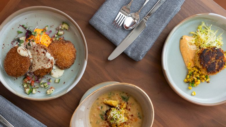 Atop view of three plates with plated dishes: croquettes or fried patties, garnishes, sauces, and a bowl of soup; cutlery and napkins nearby, wooden table.