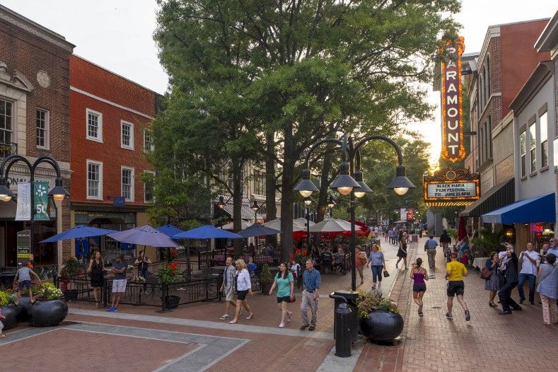 A busy pedestrian street with shops, trees, and people dining at outdoor tables by the cinema sign on the right.
