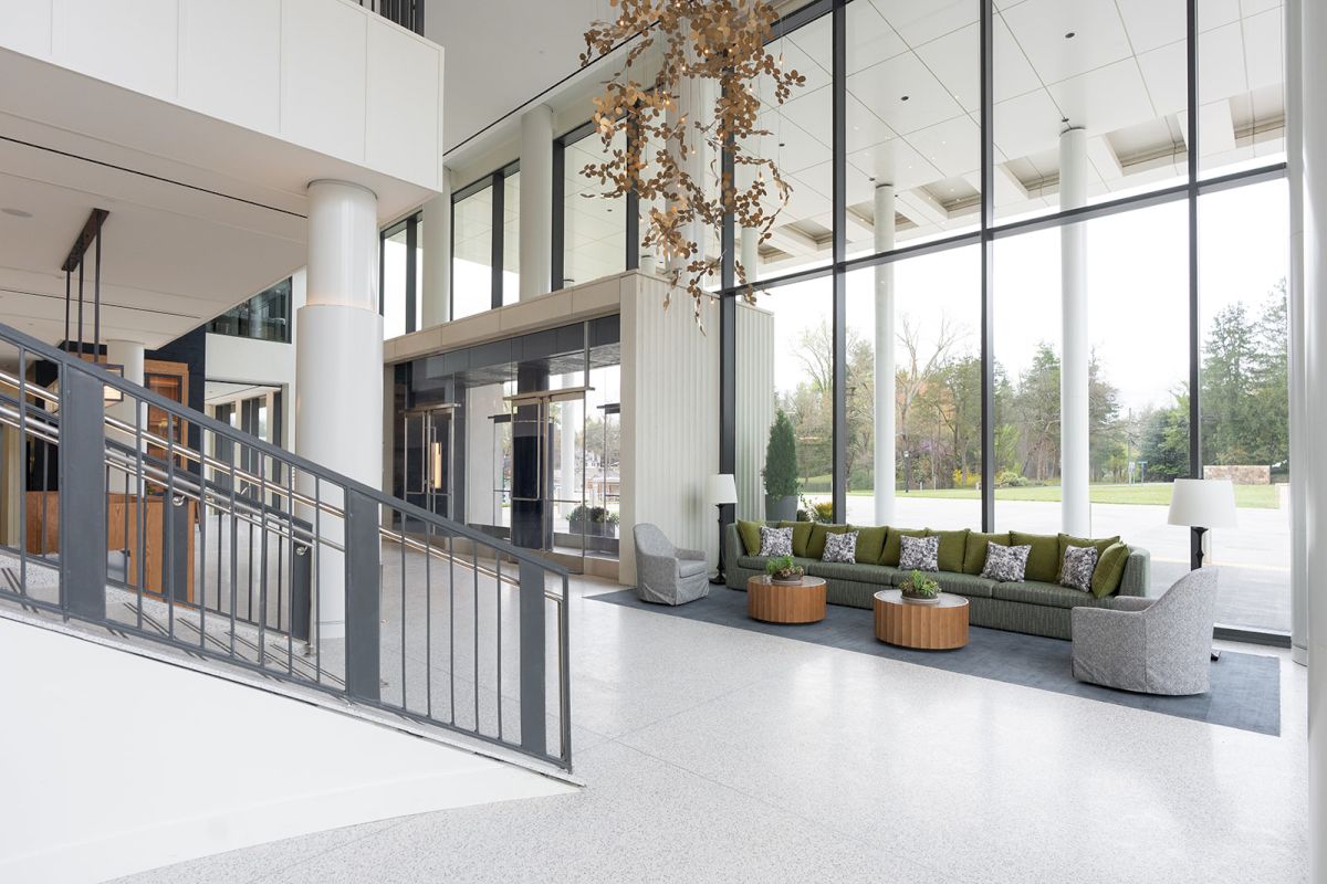 A modern lobby with large glass walls, a staircase, a gray sofa seating area, potted trees, and natural light streaming in.