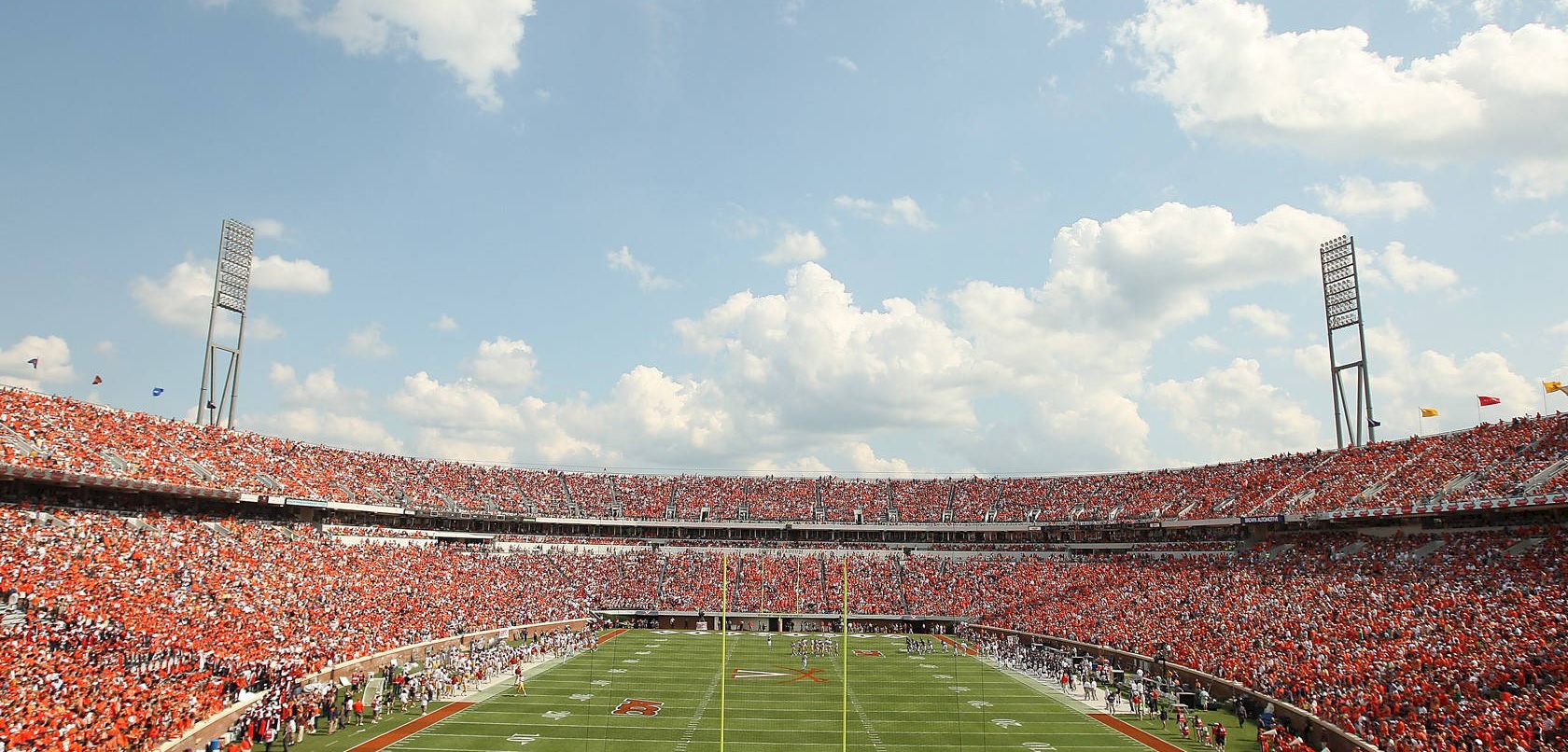 A packed football stadium with players on the field and a bright, sunny sky above, full of cheering fans in red seats.