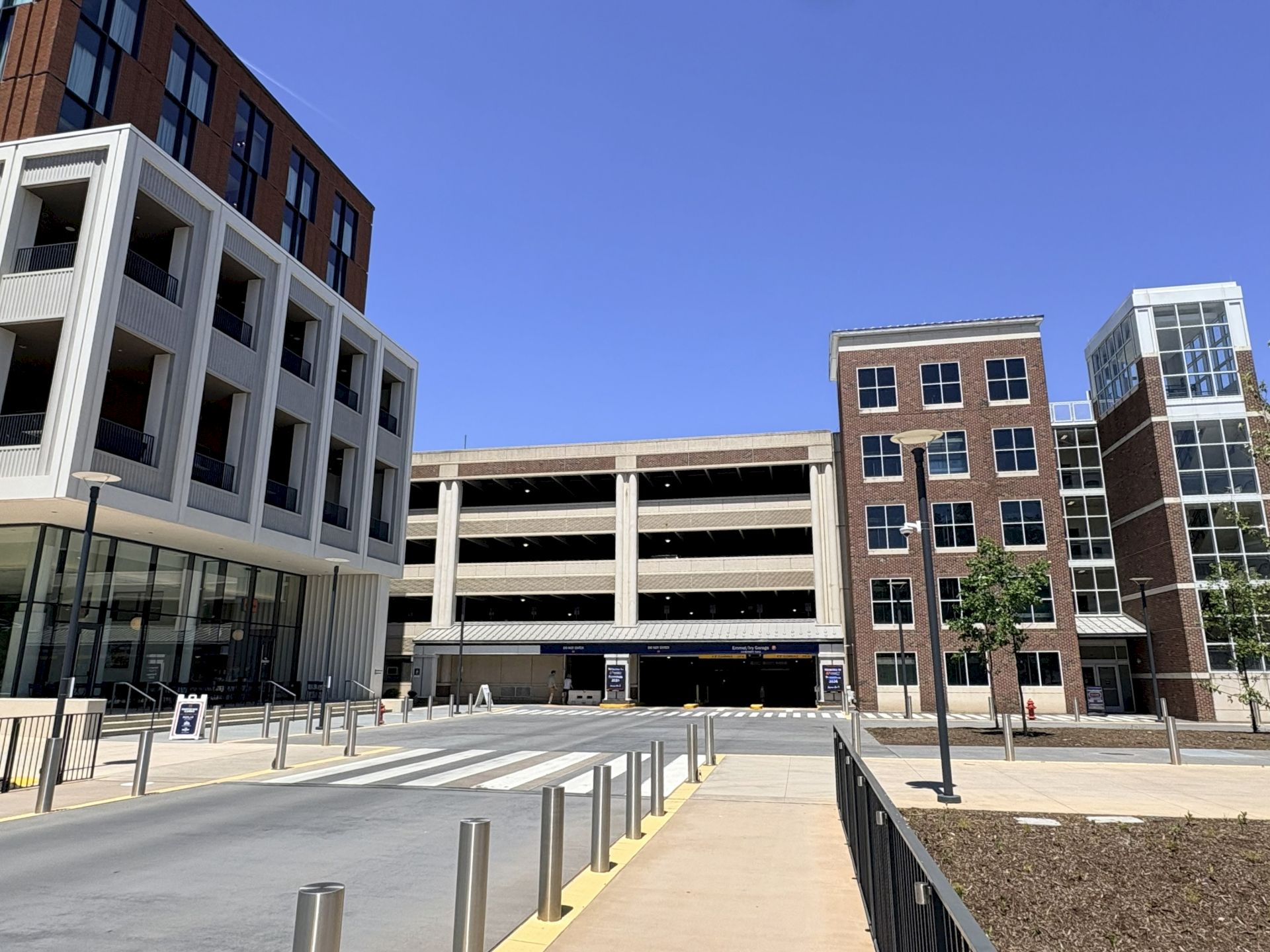 A modern urban scene with a multi-story parking garage between brick buildings, a wide sidewalk, bollards, and a clear blue sky ending.