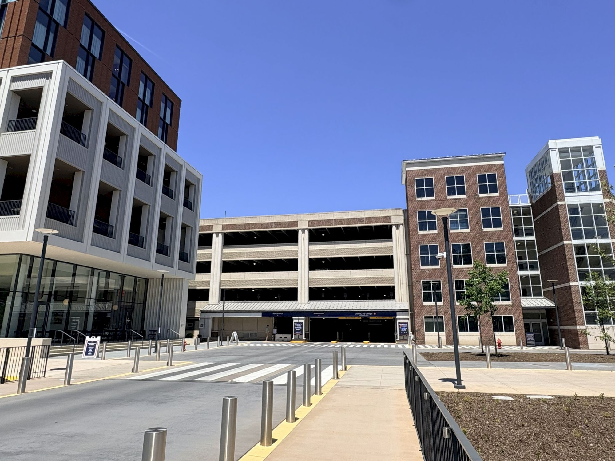 A modern urban scene with a multi-story parking garage between brick buildings, a wide sidewalk, bollards, and a clear blue sky ending.