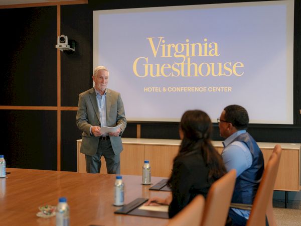 A speaker presents in a meeting room for Virginia Guesthouse Hotel & Conference Center, with attendees seated in front of a screen.
