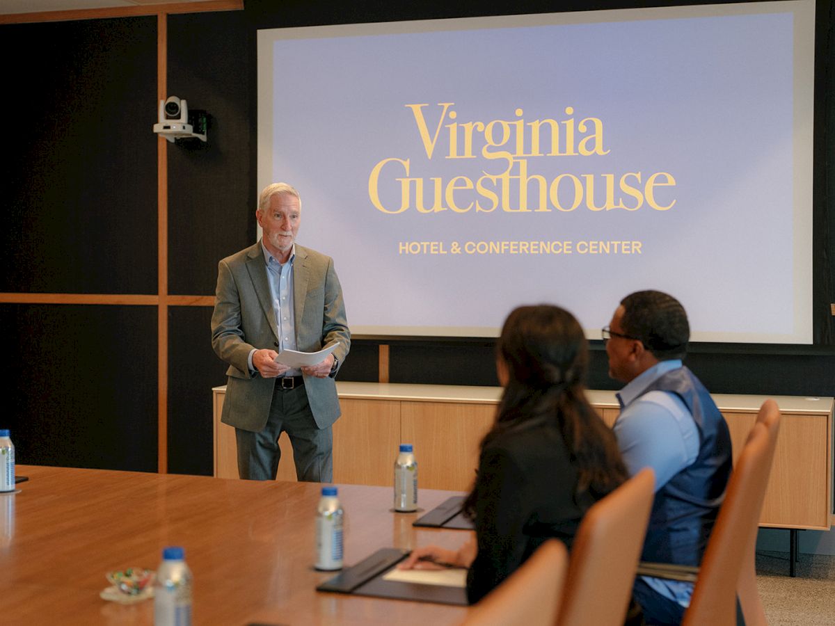 A speaker presents at a meeting room for Virginia Guesthouse, with attendees seated at a conference table.