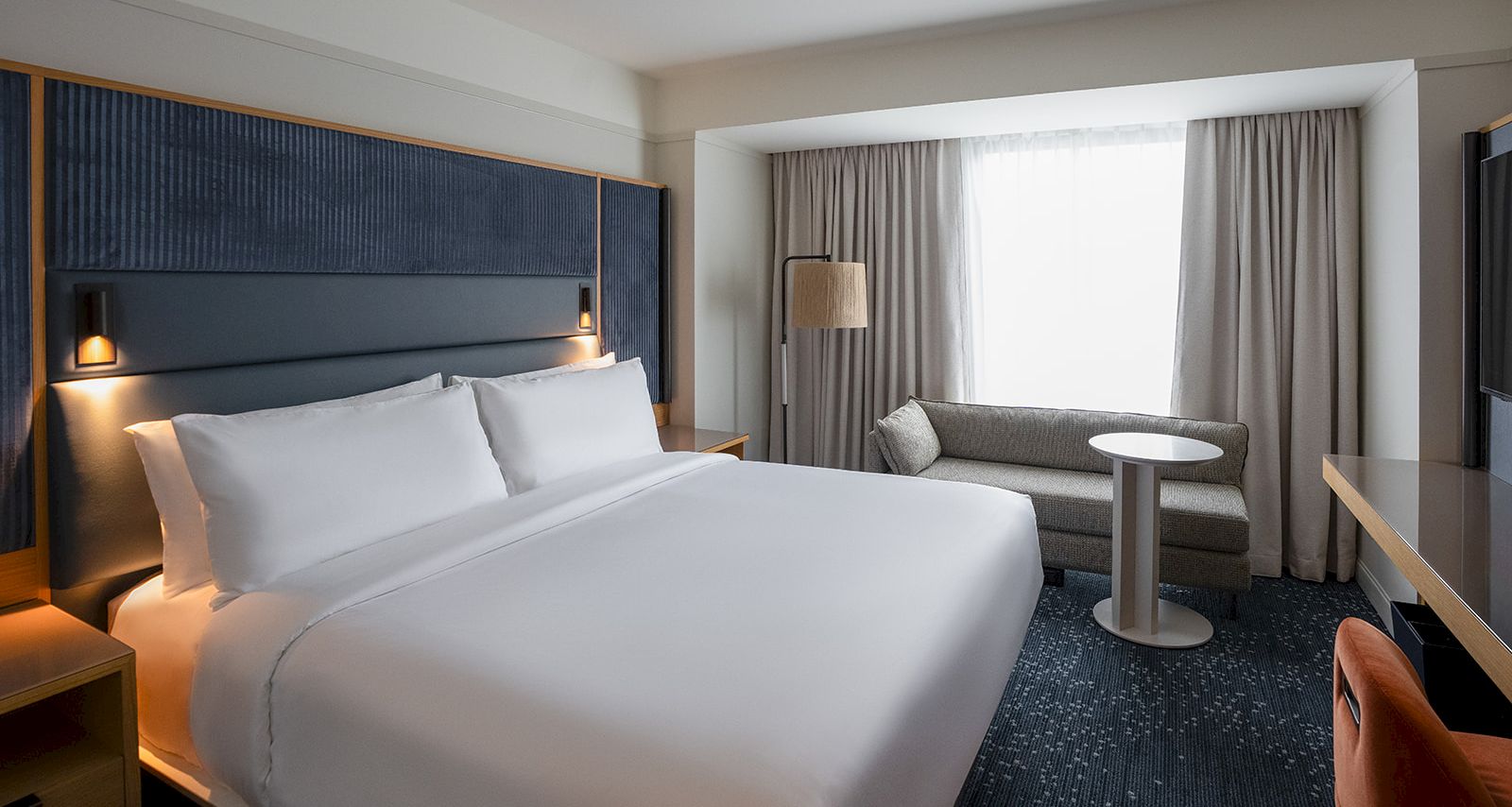 A modern hotel room with a neatly made bed, bedside lamps, curtains, and a small seating area by a window.