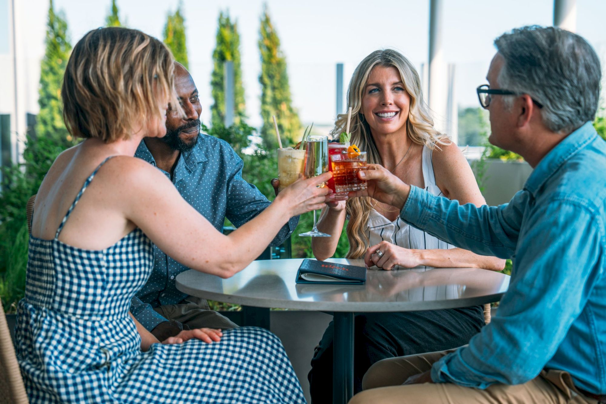 Friends toast colorful cocktails on a sunny patio, chatting with drinks held up in the air.