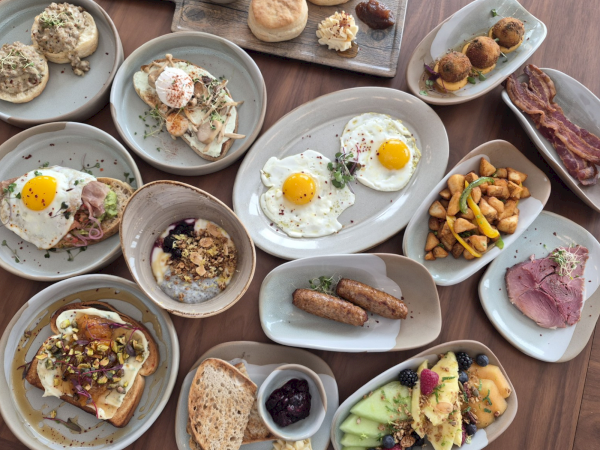 A brunch spread of fried eggs, toast, pastries, sausages, bacon, potatoes, and fruit on shared plates.