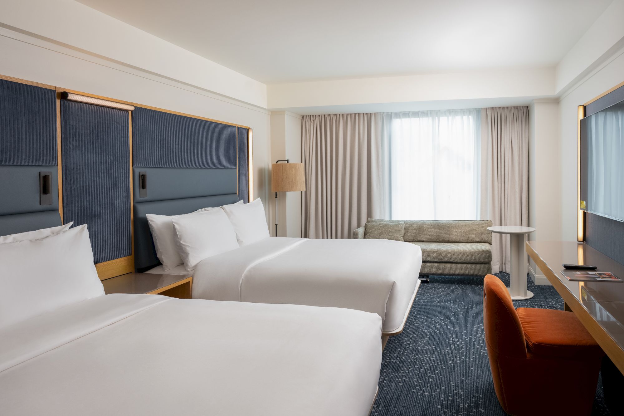 A modern hotel room with two beds, a sofa, bedside lamps, and large curtains near a window.