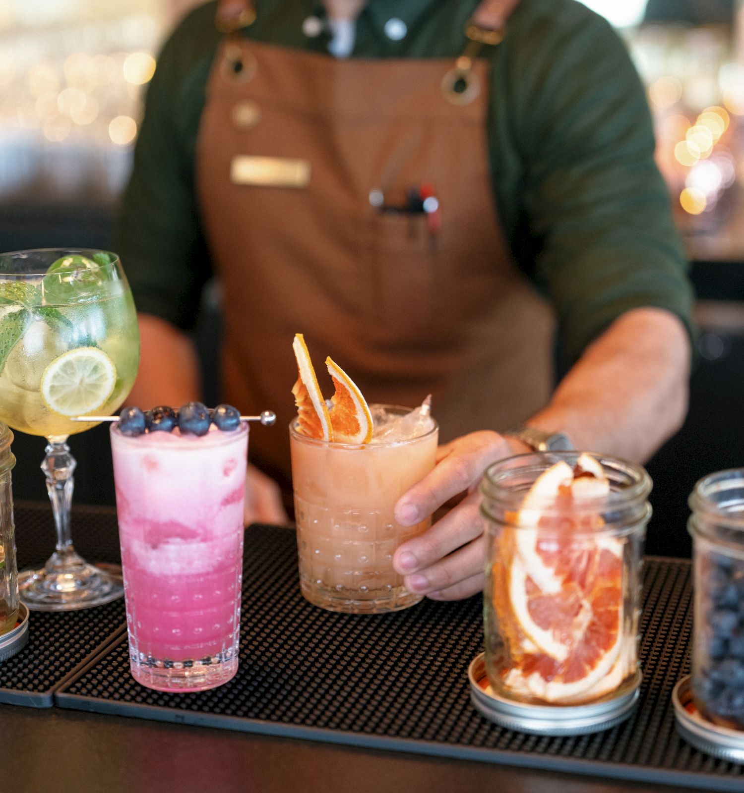 A bartender in an apron serves cocktails at a bar, with pink and amber drinks garnished with fruit and olives.