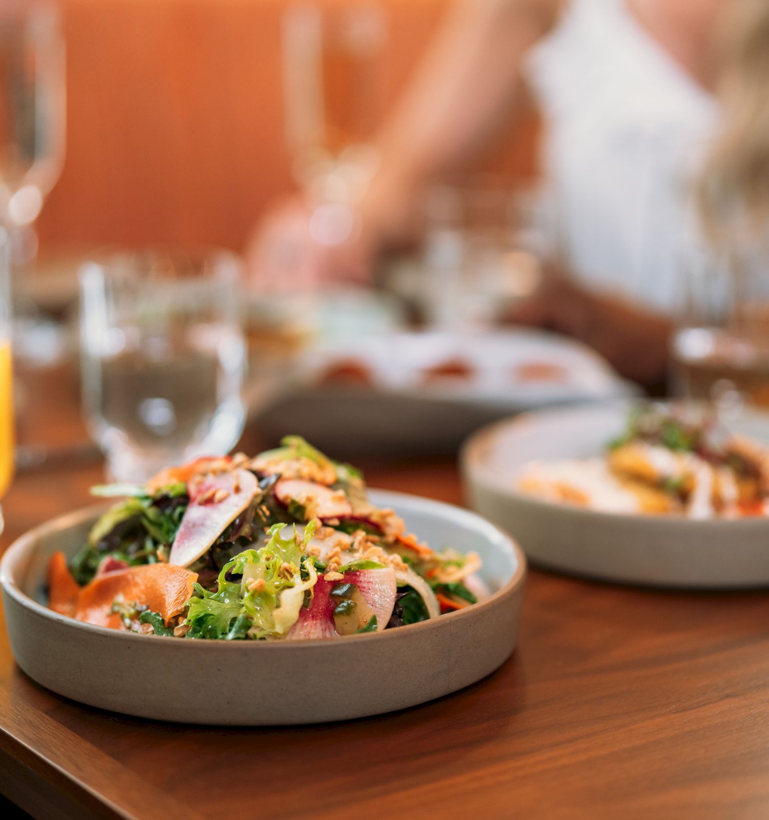 A fresh salad in a bowl with a glass of orange juice on a table, with another dish in the background.