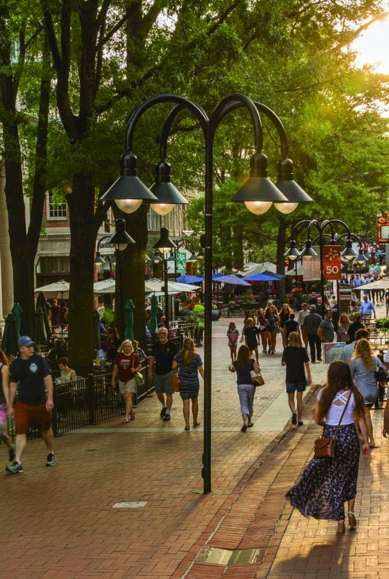 Bustling city street with shoppers under trees, outdoor cafes, storefronts, and a &ldquo;rockpaperscissors&rdquo; sign in warm sunlight.