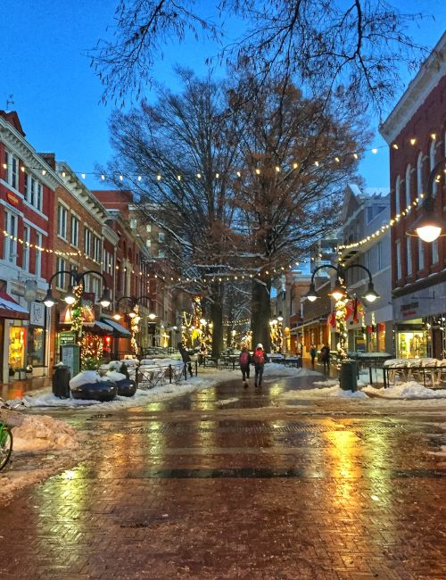 Snowy downtown street with shops, string lights, and streetlamps lining a wet road on a clear blue evening.