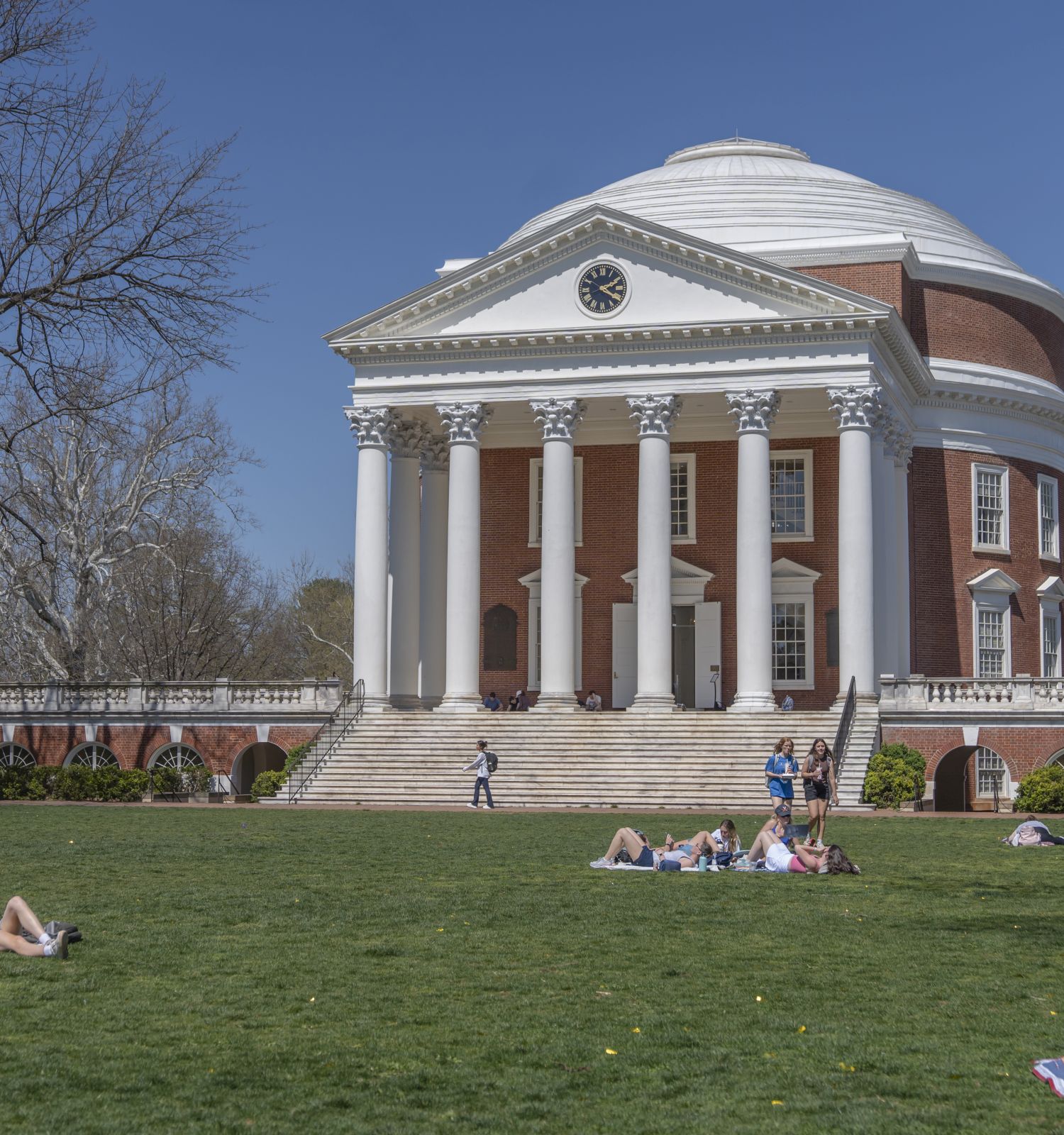 A red-brick neoclassical building with white columns and a dome clock, with students relaxing on the green lawn under blue sky.
