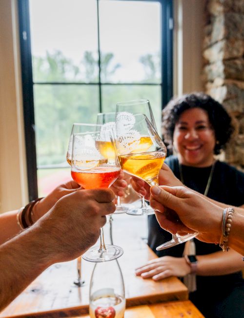 Friends clink craft beer glasses with amber and orange drinks in a cozy brewery-like room by a window.