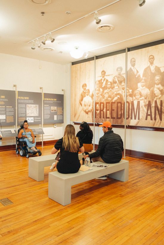 A museum exhibit room with people gathered around displays, featuring a large wall banner reading &ldquo;Freedom and Education.&rdquo;