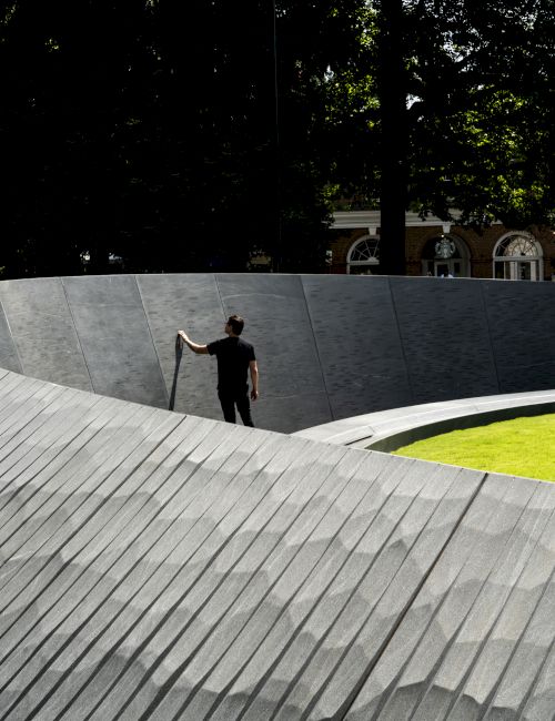 A man walks along a curved metal memorial wall in a park, with trees, grass, and a Starbucks-style building in the background.