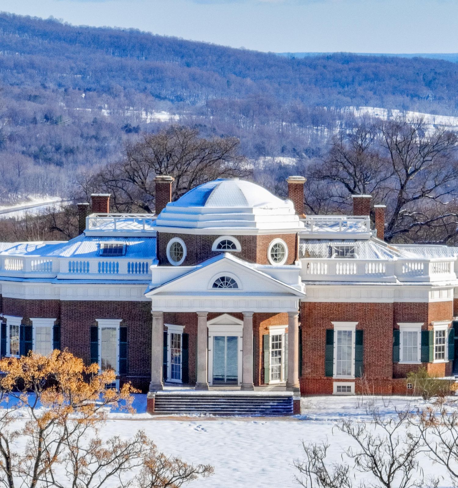 Snowy brick mansion with white columns and a blue-tinted glass roof, overlooking winter hills.