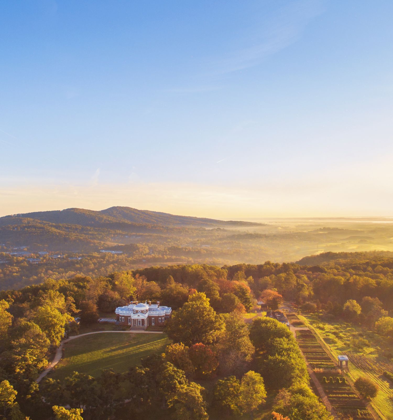 Aerial sunrise over a lush valley with forests, rolling hills, farmland, and a grand house in front.